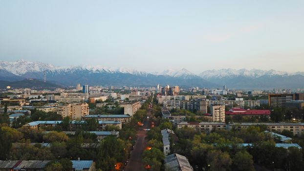 View of the snowy mountains of the TRANS-ili Alatau and the city of Almaty. Clear blue sky, lights are on in the city, cars are driving on the road. Courtyards and streets of the city are illuminated