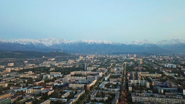 View of the snowy mountains of the TRANS-ili Alatau and the city of Almaty. Clear blue sky, lights are on in the city, cars are driving on the road. Courtyards and streets of the city are illuminated