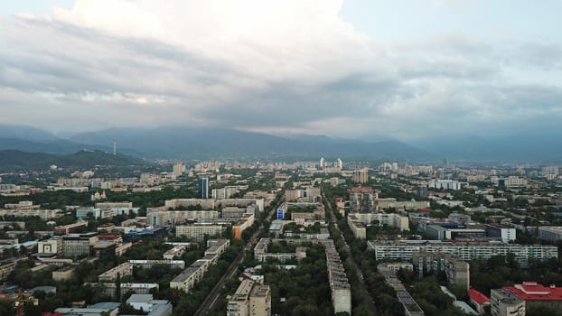 Huge clouds over the city of Almaty. Clouds hang near the mountains. Sunset. You can see different buildings, mosques, churches, and parks. Everything is illuminated by the red rays of the sun.