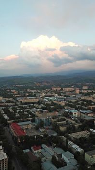 Huge clouds over the city of Almaty. Clouds hang near the mountains. Sunset. You can see different buildings, mosques, churches, and parks. Everything is illuminated by the red rays of the sun.