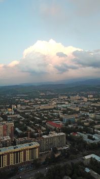 Huge clouds over the city of Almaty. Clouds hang near the mountains. Sunset. You can see different buildings, mosques, churches, and parks. Everything is illuminated by the red rays of the sun.