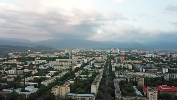 Huge clouds over the city of Almaty. Clouds hang near the mountains. Sunset. You can see different buildings, mosques, churches, and parks. Everything is illuminated by the red rays of the sun.