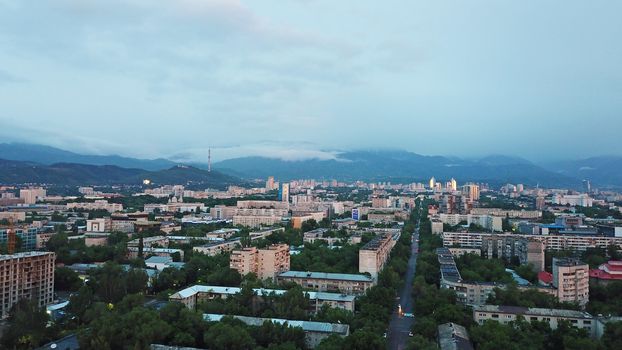 Clouds over the mountains and the city of Almaty at sunset. A lot of green trees, cars driving on the road, high mountains can be seen in the distance. On the hill stands the Kok Tobe TV tower.