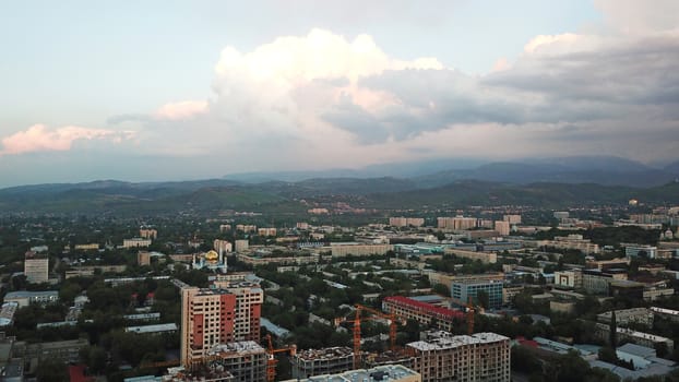 Huge clouds over the city of Almaty. Clouds hang near the mountains. Sunset. You can see different buildings, mosques, churches, and parks. Everything is illuminated by the red rays of the sun.