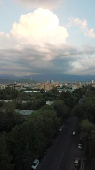 Huge clouds over the city of Almaty. Clouds hang near the mountains. Sunset. You can see different buildings, mosques, churches, and parks. Everything is illuminated by the red rays of the sun.