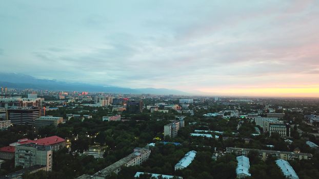 Clouds over the mountains and the city of Almaty at sunset. A lot of green trees, cars driving on the road, high mountains can be seen in the distance. On the hill stands the Kok Tobe TV tower.