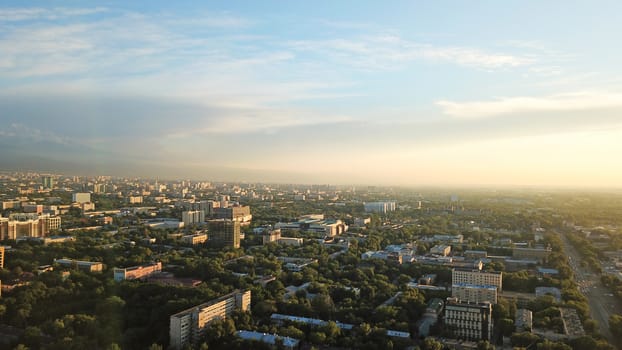 Bright color sunset over the city of Almaty. Huge clouds over the mountains and the city shimmer from bright blue to yellow and dark blue. Tall houses and green trees, cars driving on the roads.