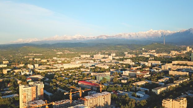 Bright color sunset over the city of Almaty. Huge clouds over the mountains and the city shimmer from bright blue to yellow and dark blue. Tall houses and green trees, cars driving on the roads.