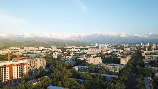 Bright color sunset over the city of Almaty. Huge clouds over the mountains and the city shimmer from bright blue to yellow and dark blue. Tall houses and green trees, cars driving on the roads.