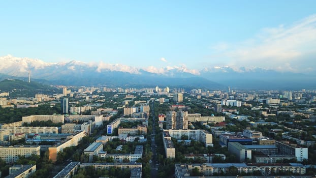 Bright color sunset over the city of Almaty. Huge clouds over the mountains and the city shimmer from bright blue to yellow and dark blue. Tall houses and green trees, cars driving on the roads.