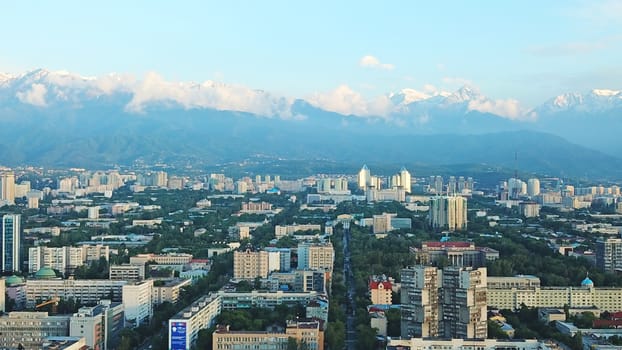 Bright color sunset over the city of Almaty. Huge clouds over the mountains and the city shimmer from bright blue to yellow and dark blue. Tall houses and green trees, cars driving on the roads.