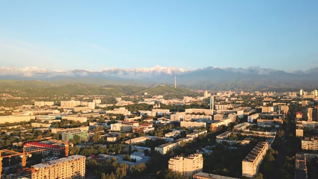 Bright color sunset over the city of Almaty. Huge clouds over the mountains and the city shimmer from bright blue to yellow and dark blue. Tall houses and green trees, cars driving on the roads.