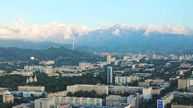 Bright color sunset over the city of Almaty. Huge clouds over the mountains and the city shimmer from bright blue to yellow and dark blue. Tall houses and green trees, cars driving on the roads.