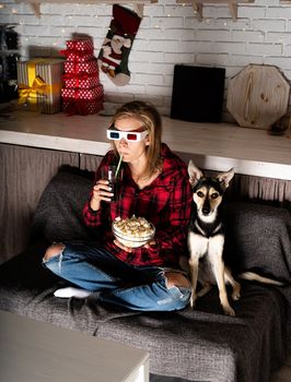 Movie night. Young woman in 3D glasses sitting on the sofa with her dog watching movies at home at night at christmas