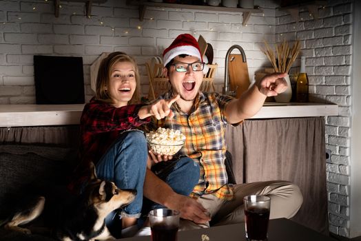 Movie night. Young couple watching movies at home at christmas pointing to the screen eating popcorn