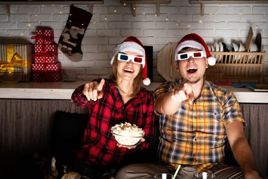 Movie night. Young couple in 3D glasses watching movies at home at christmas pointing to the screen eating popcorn