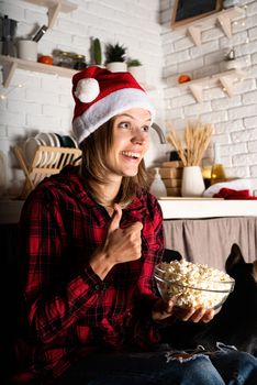 Movie night. Happy young woman in santa hat watching movies at home at christmas night eating popcorn showing thumbs up