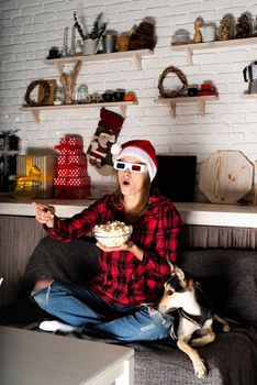 Movie night. Young woman in 3D glasses and santa hat sitting on the sofa with her dog watching movies at home at night at christmas