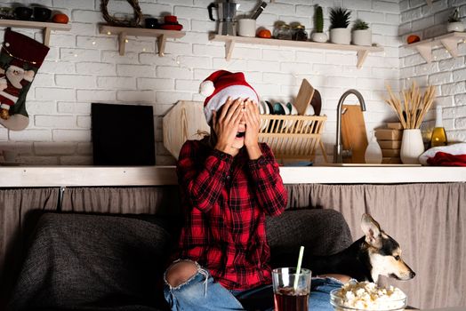 Movie night. Terrified young woman sitting on the sofa with her dog watching horror movies at home at christmas night