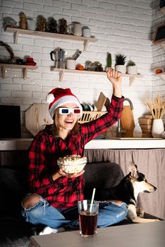 Movie night. Happy young woman in 3D glasses and santa hat sitting on the sofa with her dog watching movies at home at christmas night