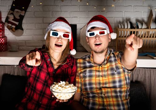 Movie night. Young couple in 3D glasses watching movies at home at christmas pointing to the screen eating popcorn