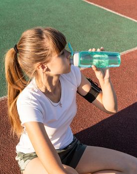 Fitness and sports. Teenage girl sitting at the stadium and drinking water after work out