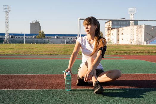 Fitness and sports. Teenage girl sitting at the stadium and drinking water after work out