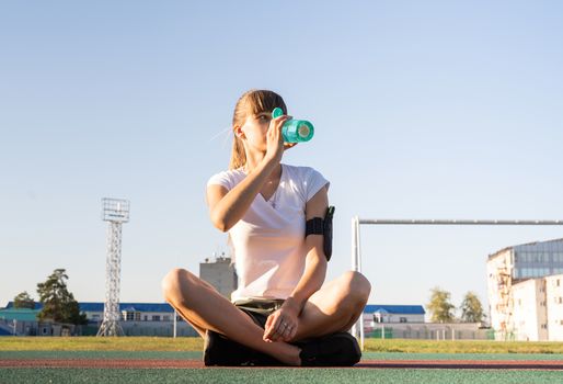 Fitness and sports. Teenage girl sitting at the stadium and drinking water after work out