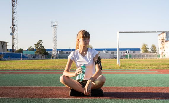 Fitness and sports. Teenage girl sitting at the stadium and drinking water after work out