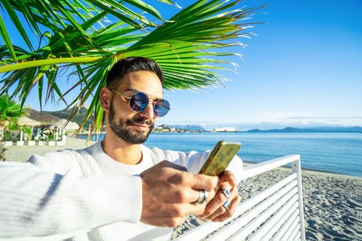 Handsome Caucasian bearded man with round sunglasses using smartphone under a palm on the beach - Stylish male person texting on phone messaging his friends and sharing his alone winter sea vacation