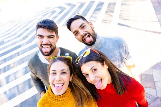 Two Caucasian couple looking at the camera from the bottom smiling and making mouths open faces sticking out their tongues - Friends having fun outdoor doing selfie with camera on top - Focus on men