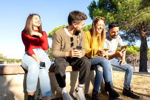 Two young beautiful heterosexual couple laughing each other using smartphone to chatting online - Happy millennial group of people having fun outdoor looking at shared contents on social network