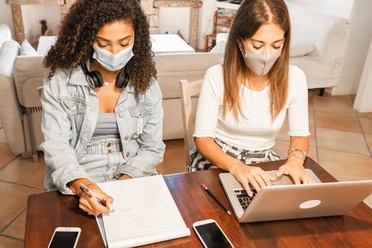Two young mixed race beautiful women working from home sitting on a wooden table in elegant living room wearing Coronavirus safe protection mask - College female students using laptop to study online