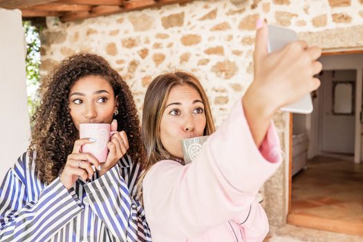 LGBT female couple making a self portrait wearing pajama in vacation home - Cute blonde Caucasian woman take a selfie with her black Hispanic curly girlfriend in the morning drinking from a tea cup
