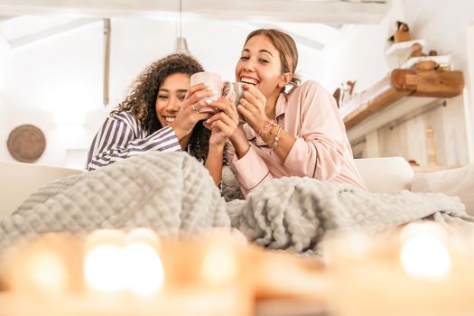 LGBT female couple having fun under a blanket on the sofa - Cute blonde Caucasian woman drink cup of tea in living room laughing with her black Hispanic curly girlfriend just woke up in the morning