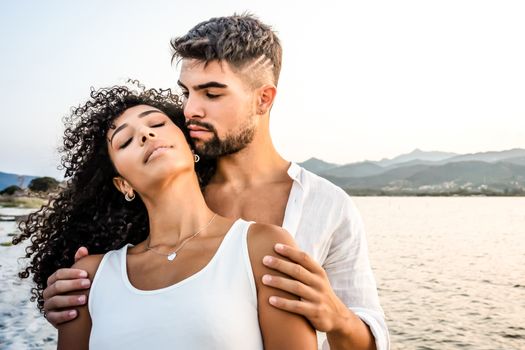 Heterosexual mixed race couple in romance scene at sunset with vintage photo effect filter - Handsome Caucasian guy embracing from back his Hispanic girlfriend with closed eyes - Focus on curly hair