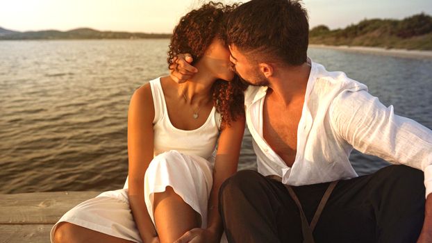 Romance scene of heterosexual mixed race couple of lovers kissing with passion on a wooden pier at sunset - Romantic handsome stylish young man flirting embracing her Hispanic curly girlfriend