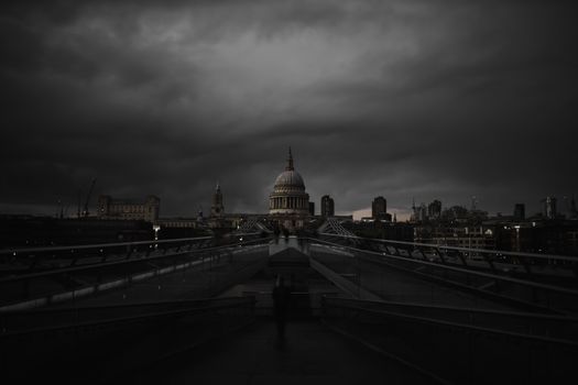 Millennium Bridge ramp with the St Paul Cathedral in the distance below creepy-looking cloudy sky. Dark gray skyline above British cathedral during dusk. Dark London cityscapes