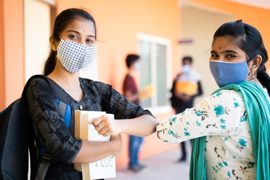 Concept of college reopen, two girls in medical mask busy in talking and moving away by greeting with elbow bump with students in background during coronavirus or covid-19 pandemic