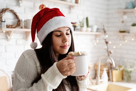 Christmas and New Year. Brunette woman in santa hat holding a cup of marshmallow cocoa in the kitchen
