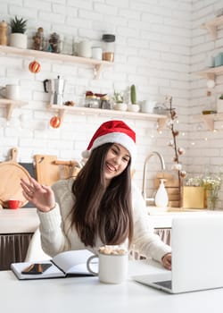 Merry Christmas and Happy New Year. Young brunette woman in santa hat chatting with friends using her laptop at the kitchen