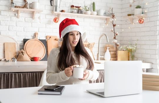 Merry Christmas and Happy New Year. Young surprised brunette woman in santa hat chatting with friends using her laptop at the kitchen