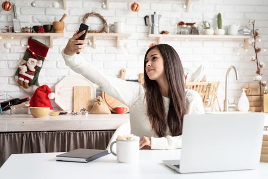 Christmas and New Year. Young brunette woman chatting with friends using her mobile phone at the kitchen