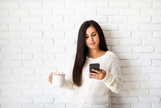 Christmas and New Year. Young brunette woman holding a cup of marshmallow cocoa and using her mobile phone on white brick wall background