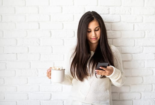 Christmas and New Year. Young brunette woman holding a cup of marshmallow cocoa and using her mobile phone on white brick wall background