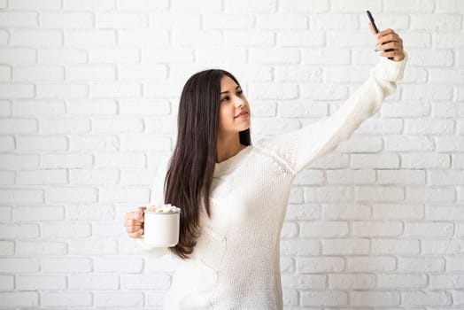 Christmas and New Year. Young brunette woman holding a cup of marshmallow cocoa and taking selfie on white brick wall background