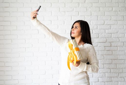 Christmas and New Year. Young brunette woman holding a gift box and taking selfie on white brick wall background