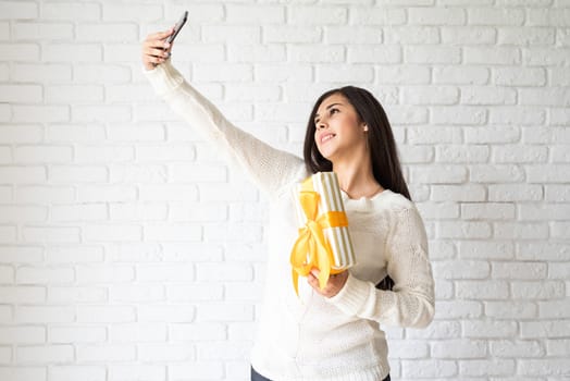 Christmas and New Year. Young brunette woman holding a gift box and taking selfie on white brick wall background