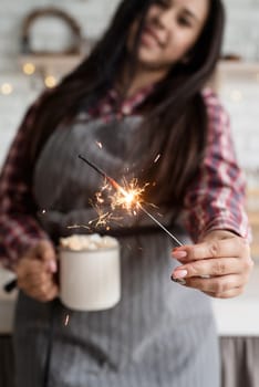 Christmas and New Year. Young smiling woman with a cup of marshmallow cocoa and a sparkler celebrating Christmas in the kitchen