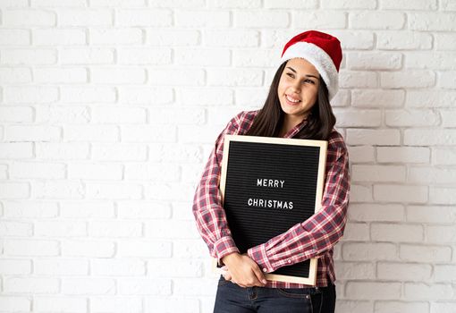 Merry Christmas and Happy New Year. Portrait of a happy beautiful latin woman with letter board with the words Merry Christmas on white brick wall background